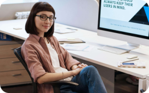 A woman with glasses sitting in front of an computer in an office space. She is wearing a beige jacket, has a short dark bowl cut hair.