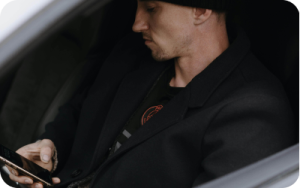 A young man sitting in a car, staring down at his smartphone. He is dressed in all black, wearing a black jacket and a black beanie. The photo is taken is through the car window.