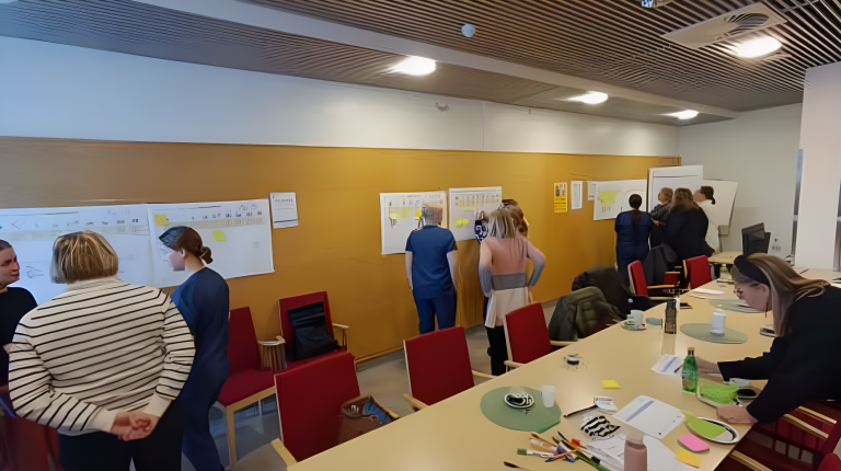 a photo of an active workshop situation. There are people in nurse uniforms and normal clothes. They are gathered in groups around big sheets of paper with sticky notes. In the foreground, there is a table with lots of office supplies like pens and sticky notes. A woman is bent over the table and is writing something.